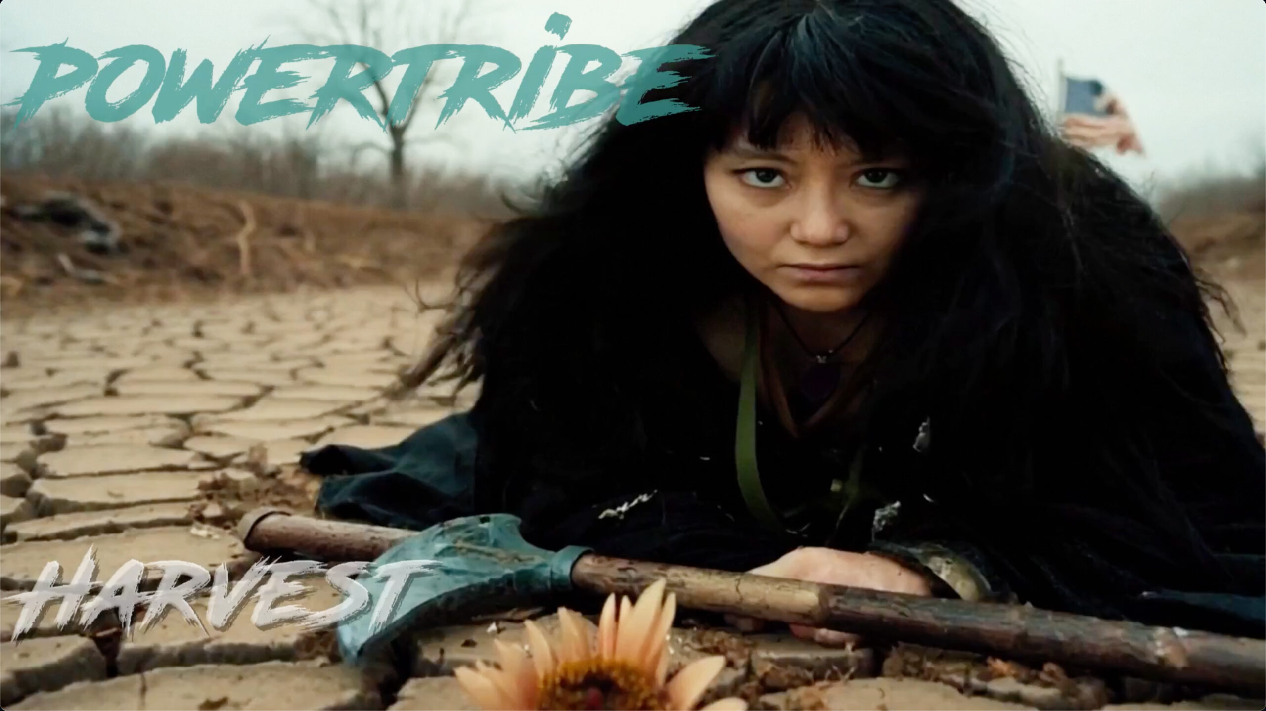 Asian woman, Missy Percifield, lies on cracked, drought-stricken earth holding a farming tool in a still from PowerTribe’s video “Harvest.”